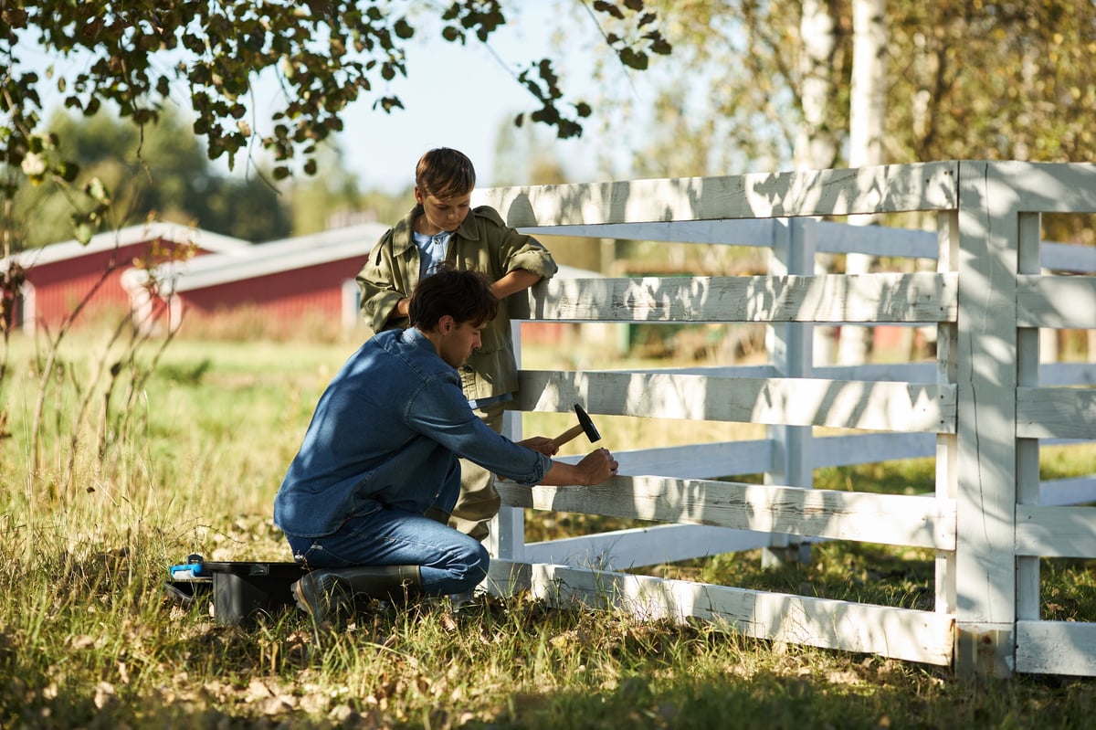Caucasian middle aged man repairing wooden fence with hammer while Caucasian boy watching outdoors on sunny day, rural setting with trees and grass in background, teamwork concept