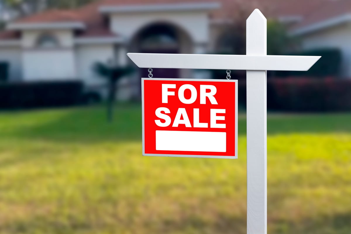 A sign stands in the grass with the words for sale on it. The sign marks a house that is available for purchase. The setting is a residential area. The time appears to be daytime with clear weather.